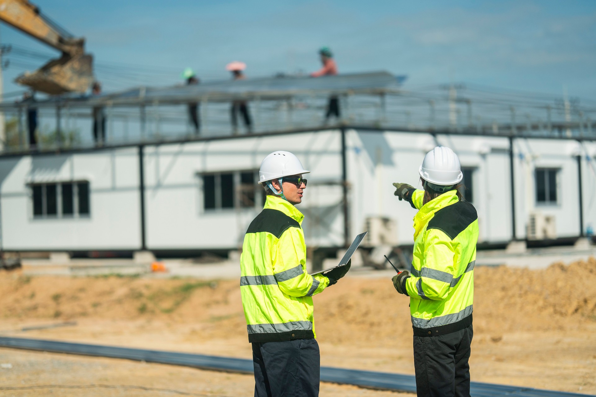 Two engineers wearing high visibility jackets and helmets discussing construction progress at a site, one pointing towards workers and machinery, emphasizing teamwork, safety, and planning in